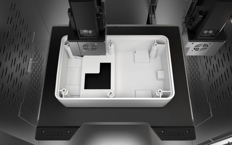 Interior view of the Intamsys Funmat Pro 310 showing a white 3D-printed box-like structure on the print bed. The print head assembly above is equipped with cooling fans and precision components. The setup emphasizes the printer's ability to handle complex builds with fine detail in an industrial setting.
