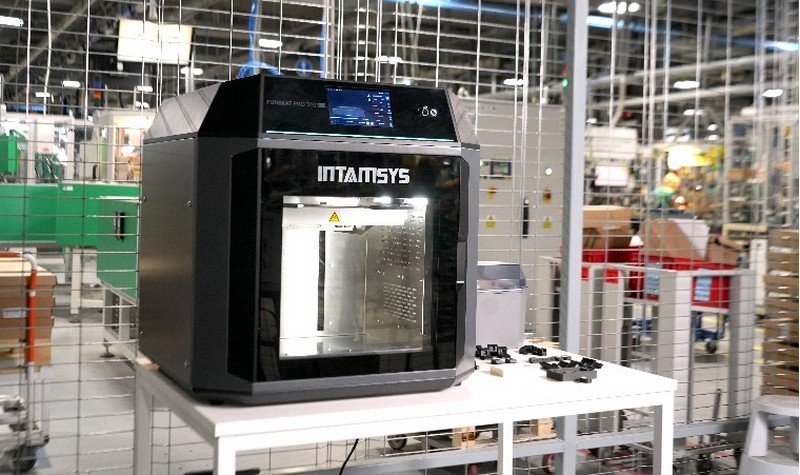 The Intamsys Funmat Pro 310 is prominently displayed in an industrial setting, resting on a white table. Its sleek, black exterior is complemented by a transparent front door that reveals the spacious printing chamber. The top section features a digital control panel with a vibrant display, allowing for precise temperature and print management. Visible branding includes the "INTAMSYS" logo on the front. The surrounding environment suggests a professional manufacturing facility with various machinery and equipment visible in the background.