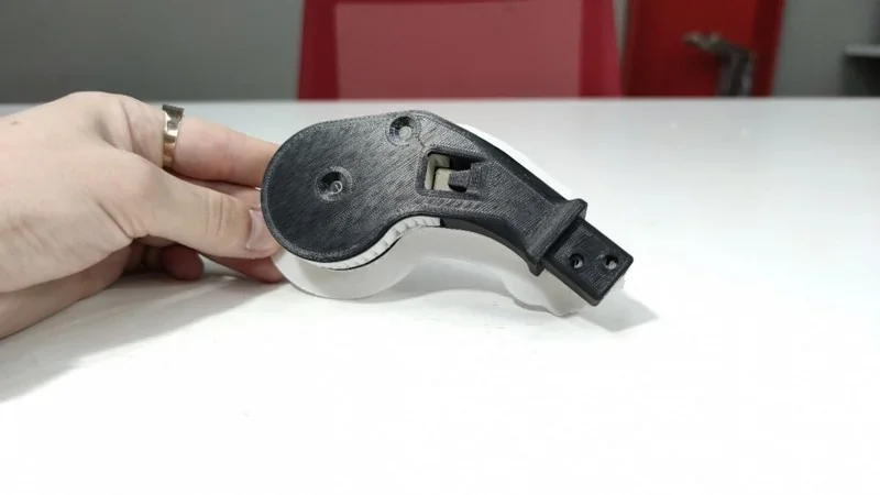 Hand holds a 3D-printed black and white component created by the Intamsys Funmat Pro 410, placed on a white table with red chairs in the background.