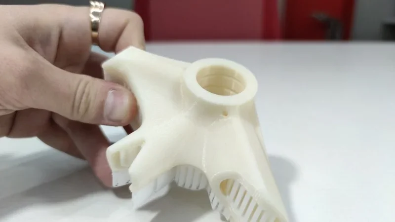 A hand holds a complex 3D-printed white part with intricate details, produced by the Intamsys Funmat Pro 410, on a white background.