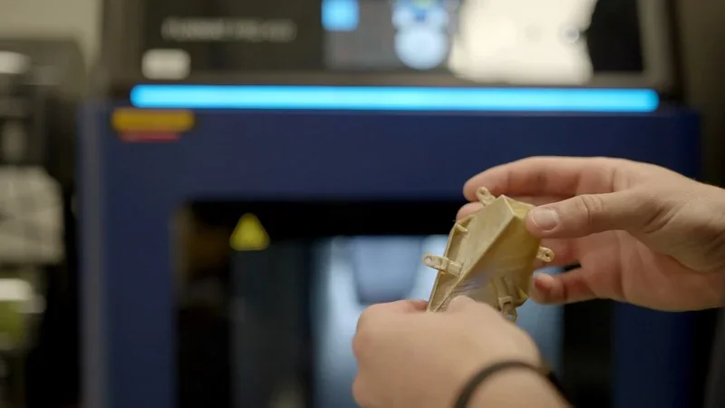 Hands holding a 3D printed part in front of an Intamsys Funmat Pro 410 printer with a blue and black interface in an industrial setting.