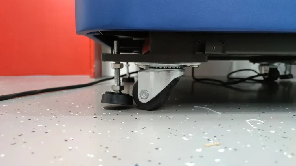 Close-up of an Intamsys Funmat Pro 410's leveling foot and caster wheel on a speckled floor, showing part of the blue base against a red wall.