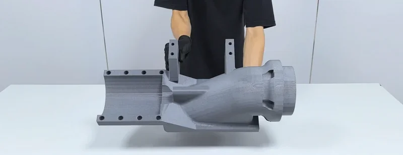 Person holds large 3D-printed industrial component created by mingda md-1000d on a table, wearing black clothing and gloves.