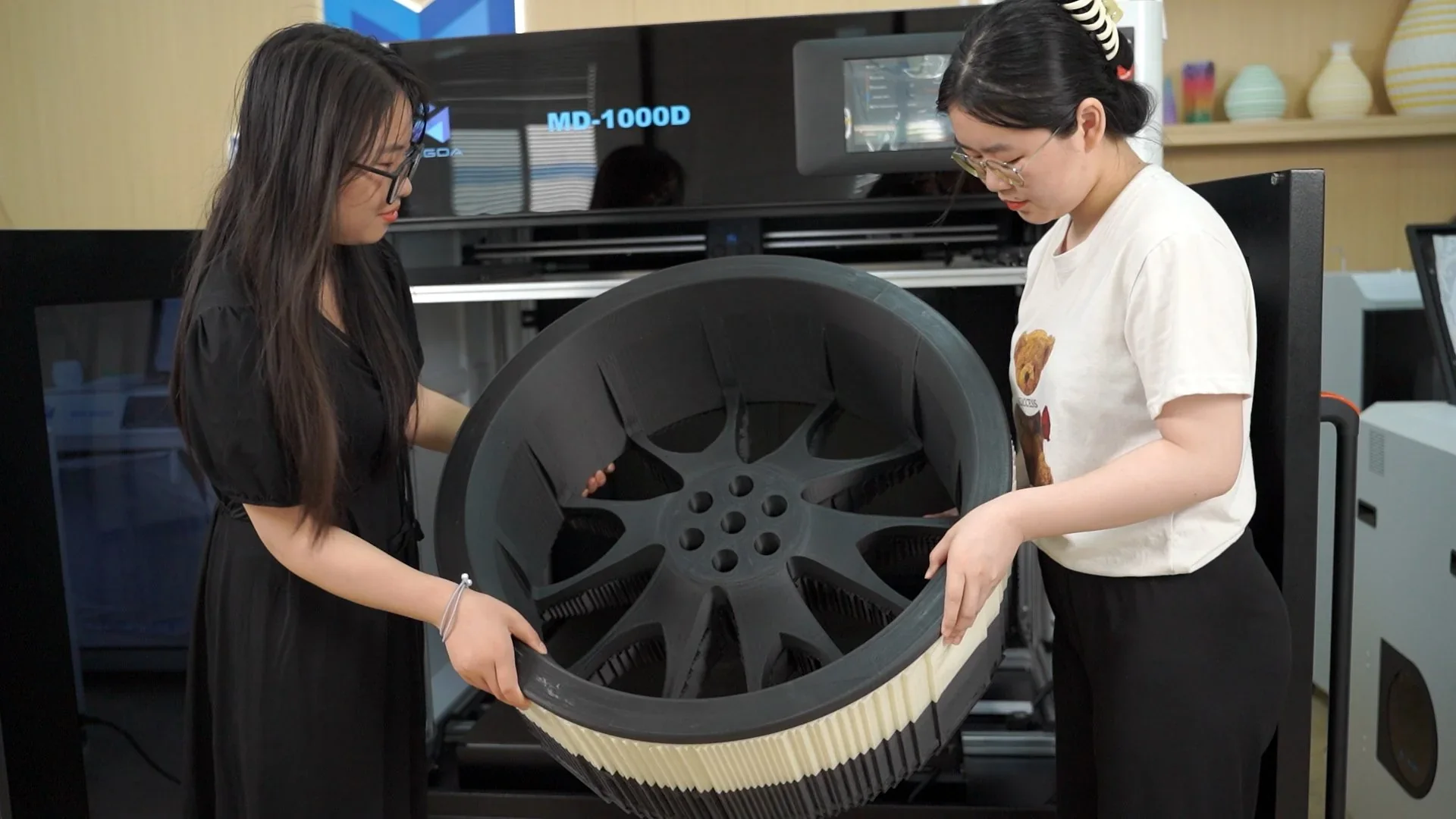 Two people hold a large 3D-printed wheel made by the Mingda MD-1000D, placed in front of the printer in an office setting.