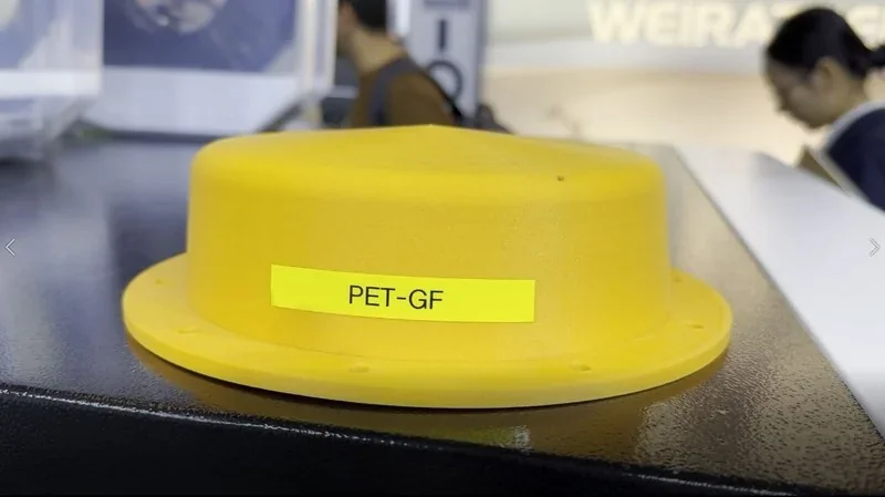 Yellow 3D-printed part by mingda md-400d labeled "PET-GF" on a black surface, displayed at a trade show with blurred people in the background.