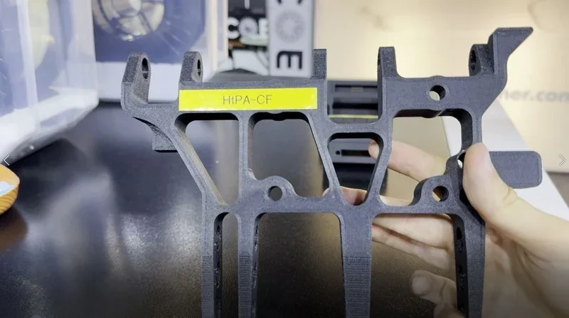 Mingda MD-400D 3D printer produces a black HIPA-CF bracket held in hand, showcasing its detailed and precise printing capabilities in a well-lit setting.