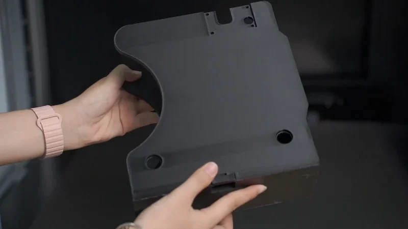 Hands holding a black 3D-printed part with holes, produced by a Mingda MD-600D printer, in an indoor setting.