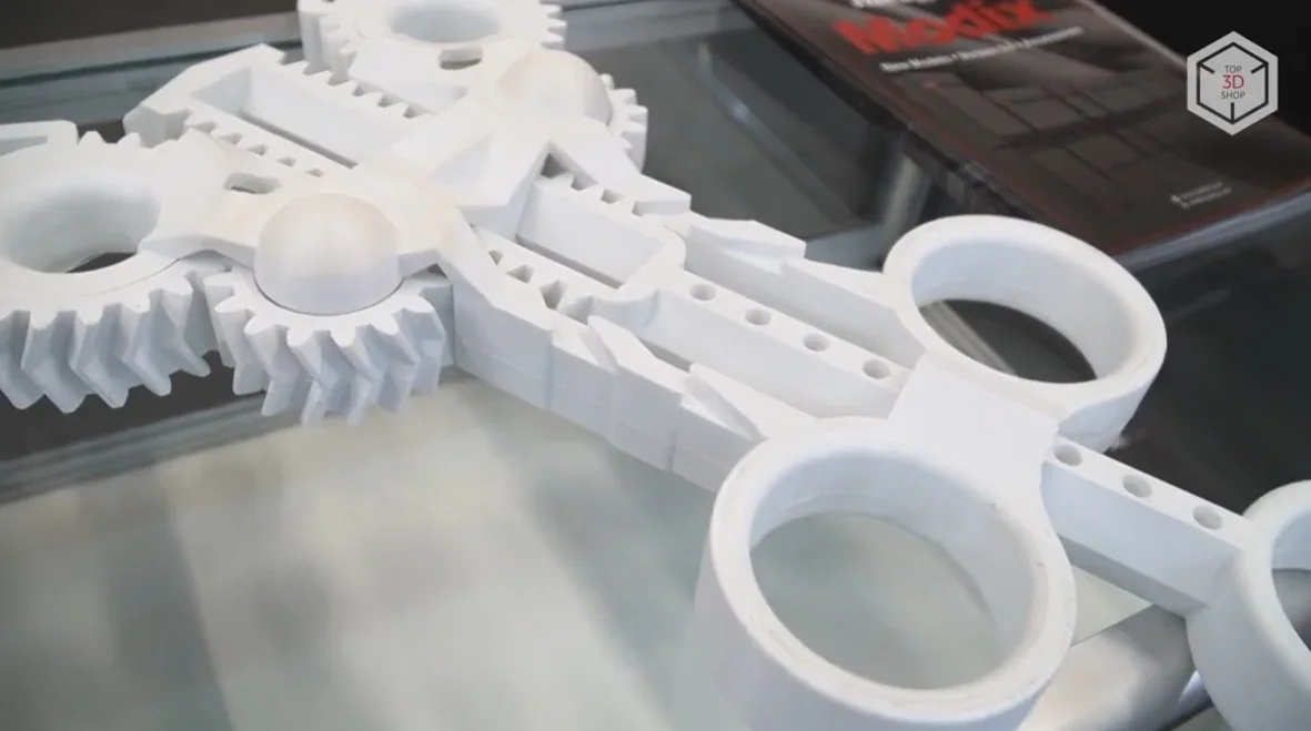 Modix Big 120X 3D printed white mechanical part with interlocking gears on display; brochure and branding visible in background.