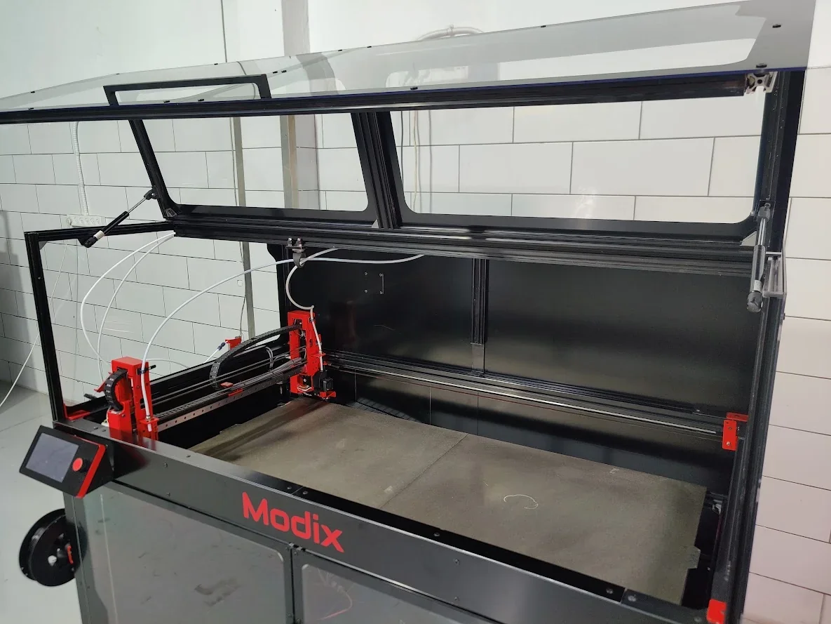 Modix Big 120x 3D printer with open transparent lid, showcasing a spacious build area, in a white-tiled room, with branding visible on the front.