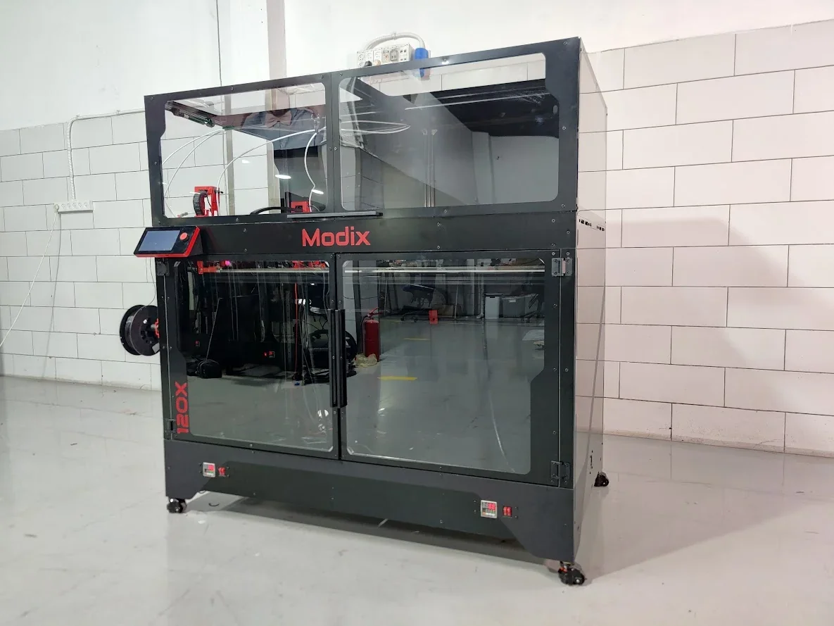 Modix Big 120x 3D printer in a white-tiled room, showcasing its large build and black casing, with visible spools and a branded display panel.