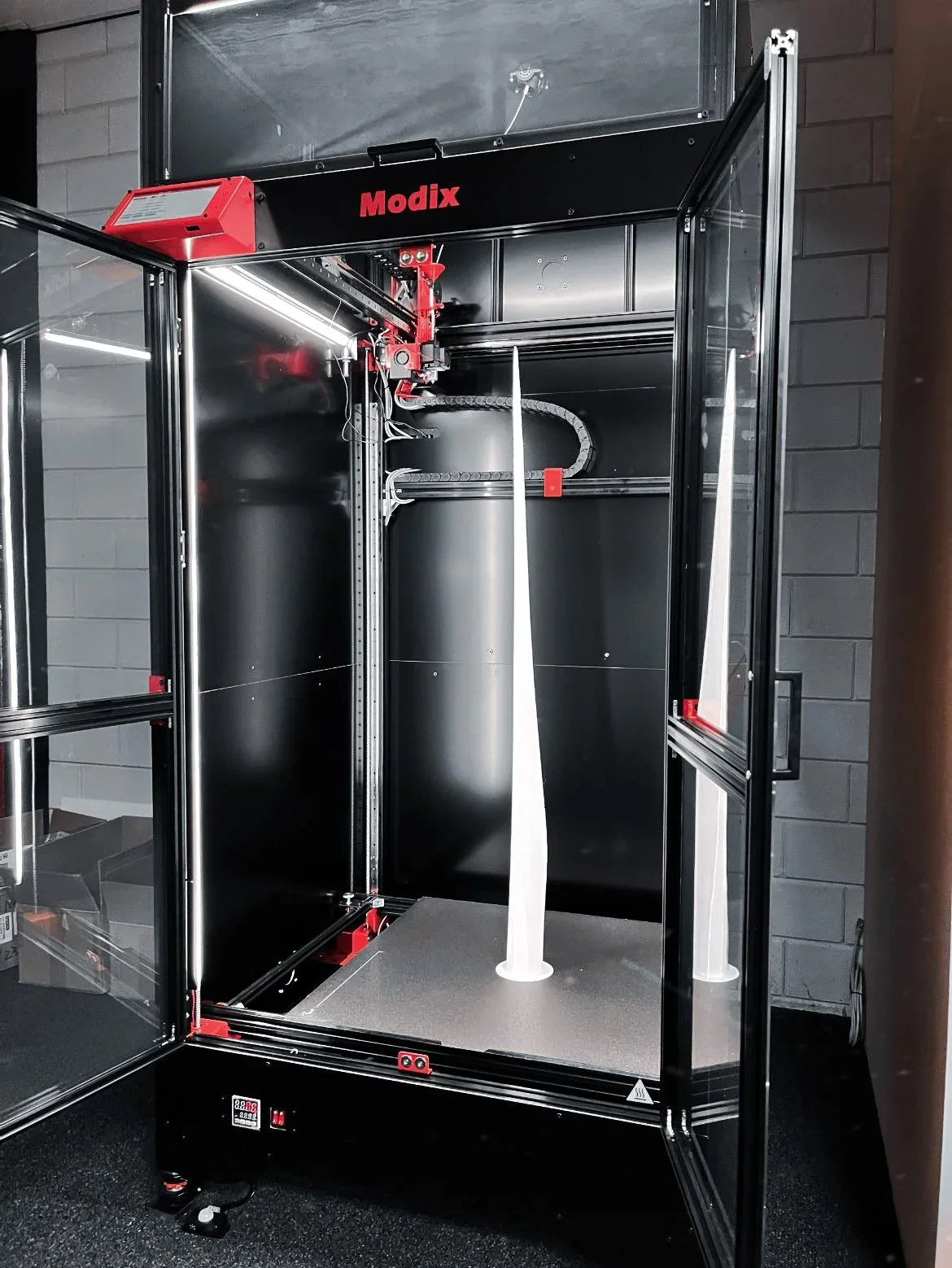 Modix Big 120Z 3D printer with an open door, showcasing a tall, white printed part inside a well-lit, industrial setting.