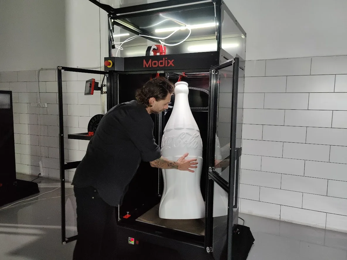 Modix Big 120Z 3D printer with an open door, showcasing a large white 3D printed bottle inside, handled by a person.