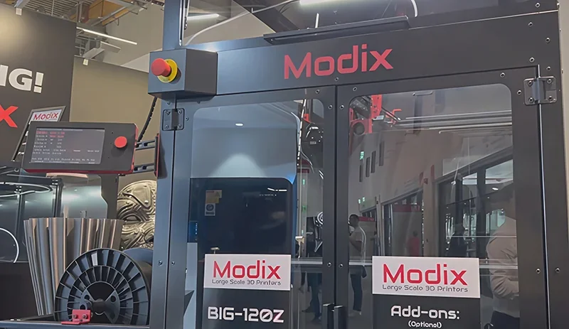 Modix Big 120Z 3D printer with control screen and branding, large filament spool, and printed parts in an industrial setting.