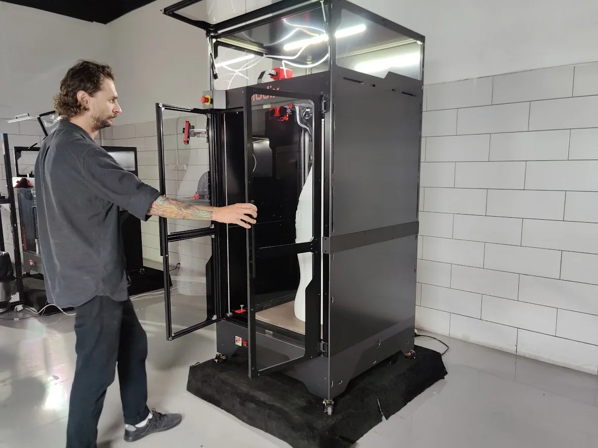 Modix Big 120Z 3D printer with a tall, dark enclosure and a user interacting. A large white print sample is visible inside. Modern workshop setting.