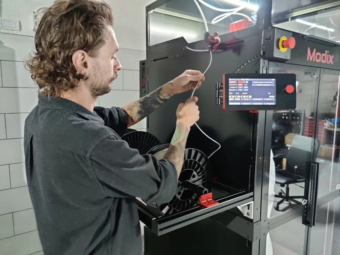 Man loading filament in Modix Big 120Z 3D printer with touchscreen and branded casing in workshop setting.