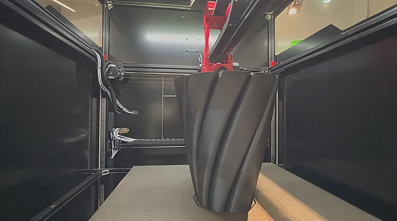 Modix Big 120Z 3D printer creating a large, black spiral vase. Interior view showcases printer components and open frame.