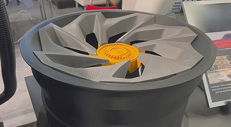 3D-printed car wheel part by Modix Big 120Z, featuring intricate blade design, displayed on a table with visible branding signage in a showroom setting.