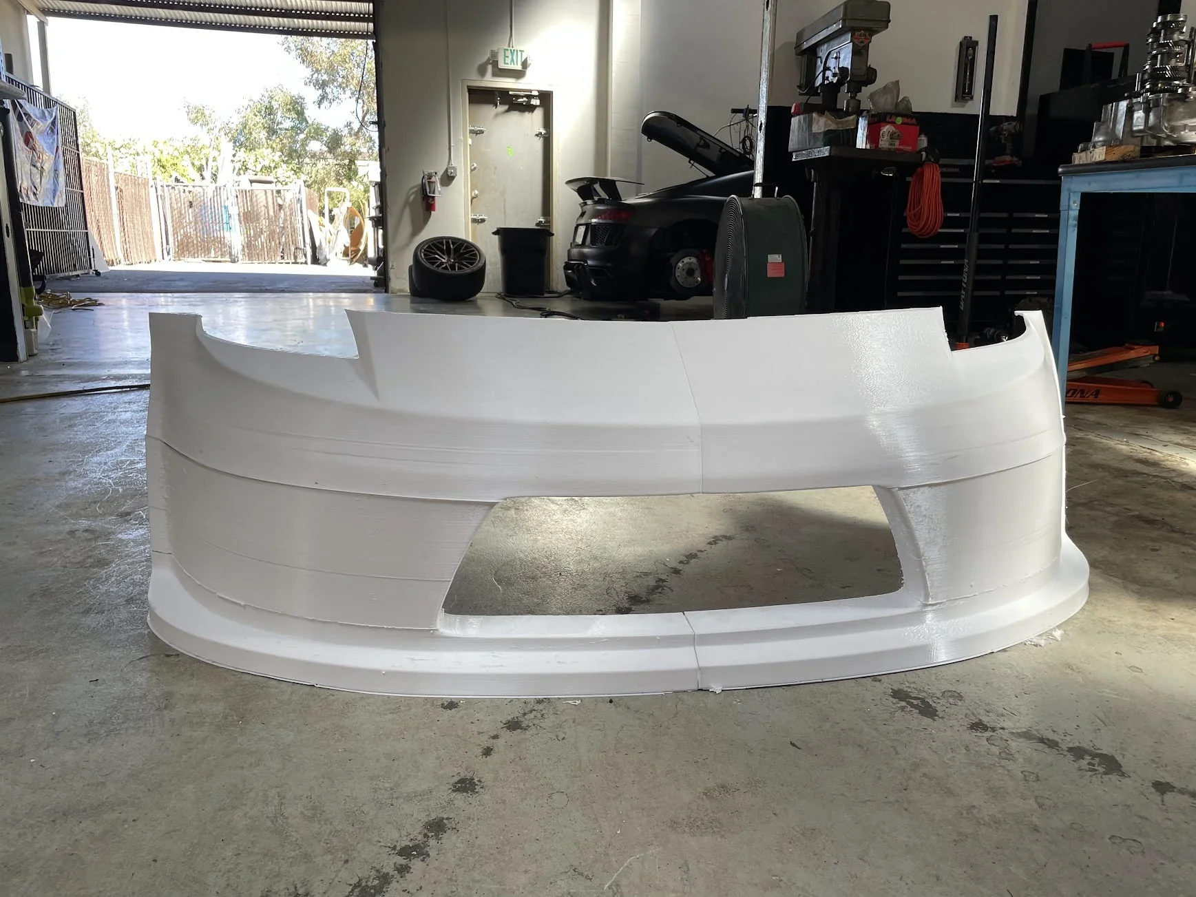 Large white 3D-printed car bumper made by Modix Big-180x in a workshop setting with tools and equipment in the background.