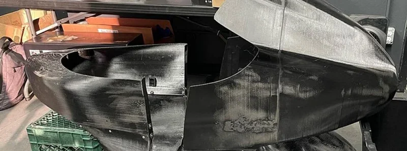 Modix Big-180x 3D printer with a large black printed part in a workshop setting, surrounded by boxes and crates.