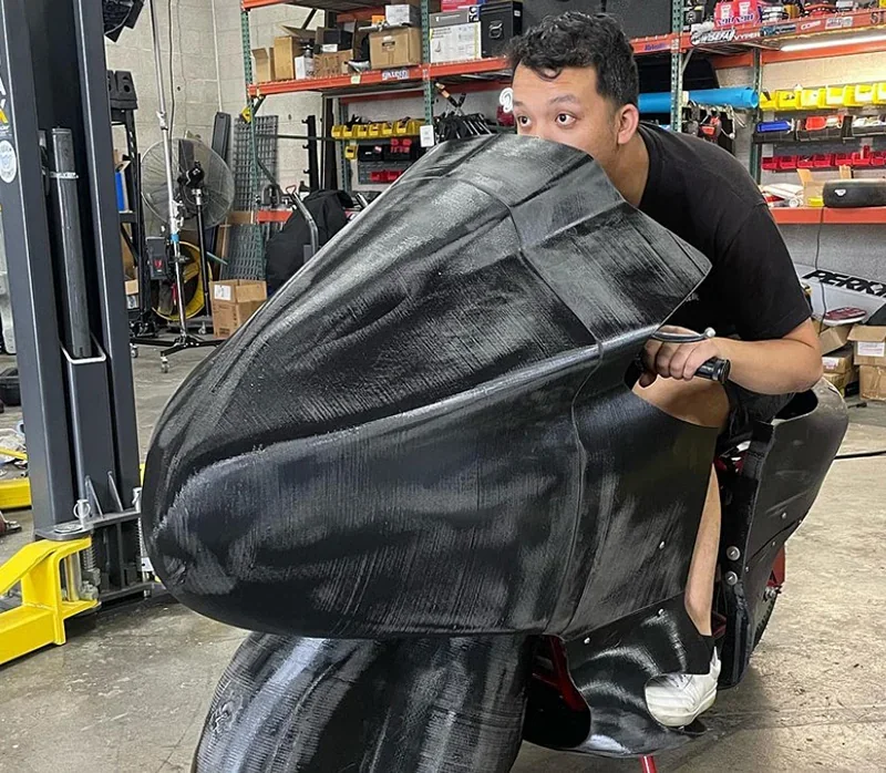 Man sitting on 3D printed motorcycle part by modix big-180x in workshop setting, showcasing large black component amid shelves and tools.