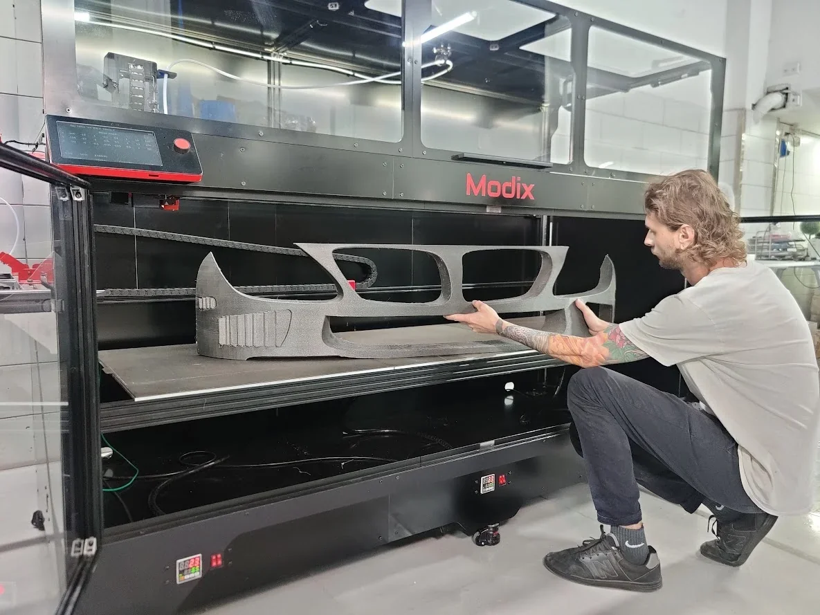 Modix Big-180x 3D printer with a man handling a large printed car bumper part in a workshop setting.