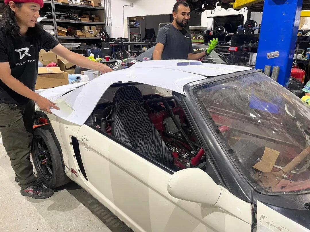 Two people work in a garage fitting a 3D-printed car roof panel, made by the Modix Big-180x, onto a white sports car amidst various tools and parts.