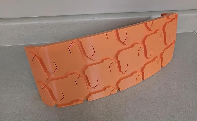 Orange 3D-printed part with geometric pattern, likely produced by Modix Big-180x, displayed on a gray surface.