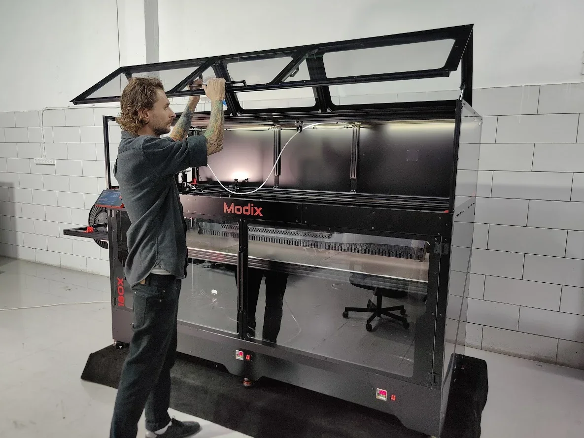 Man operating Modix Big-180X 3D printer in workshop, with transparent cover open, showing interior components and manufacturing setting.