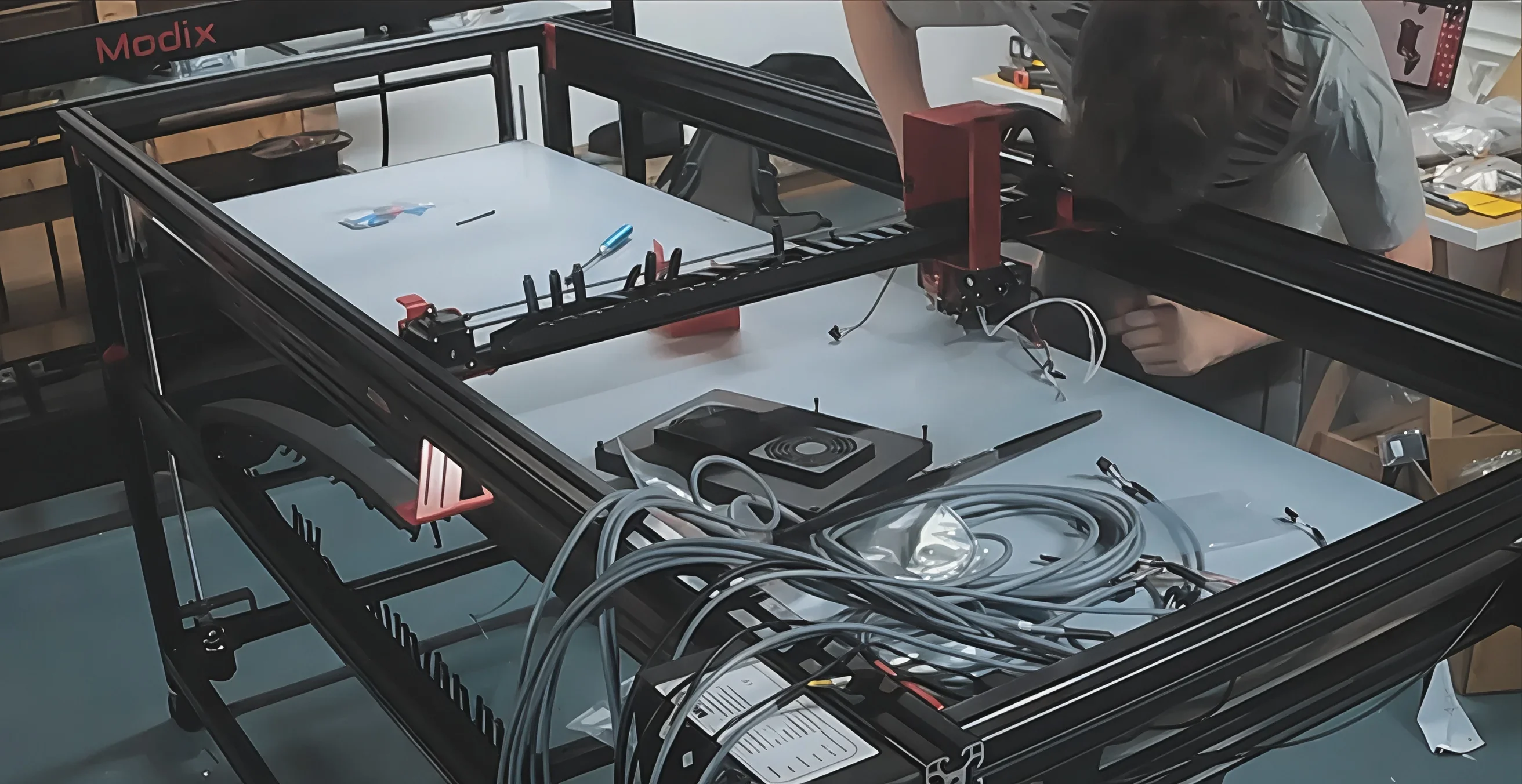 Modix Big-180x 3D printer being assembled, showing framework, wiring, and tools on a table, with visible Modix branding.