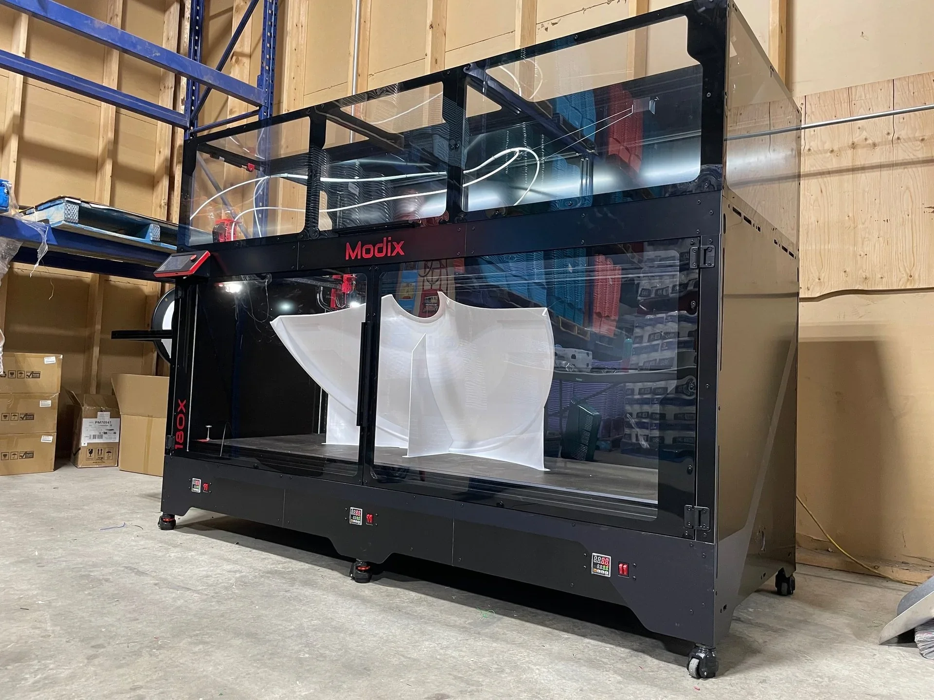 Modix Big-180X 3D printer in a workshop, printing a large white part, enclosed in a transparent case, with visible shelves and boxes around.
