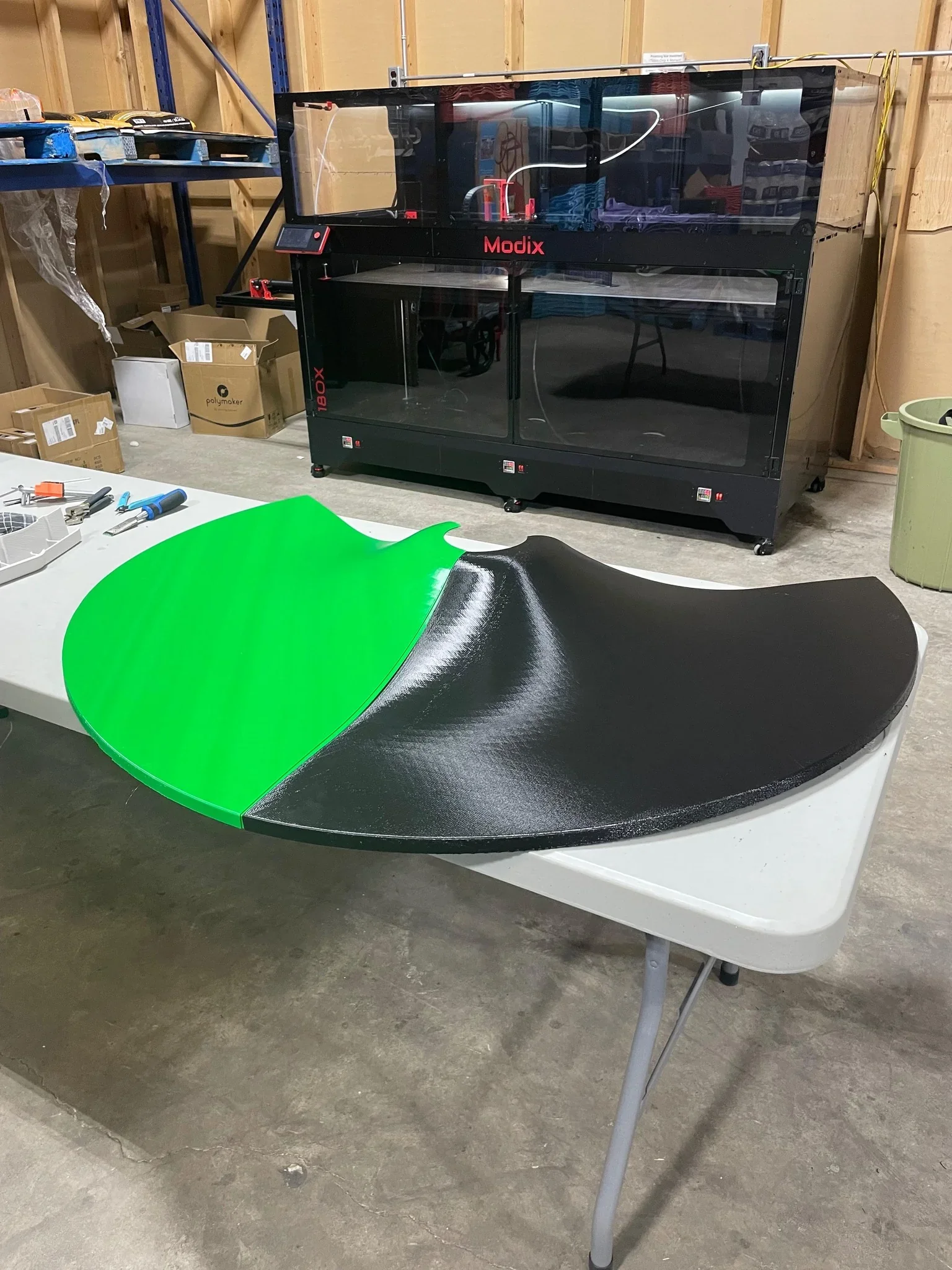 Modix Big-180X 3D printer in a workshop setting with a large, half-green, half-black printed part on a table, surrounded by tools and boxes.