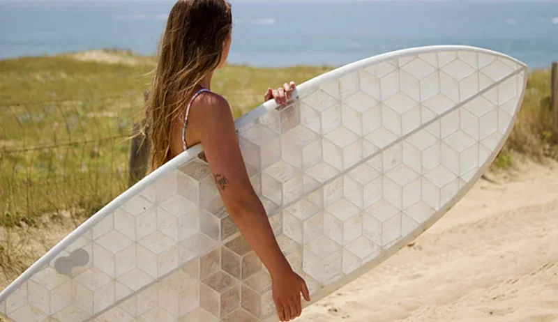 Woman holding a hexagonal-patterned 3D-printed surfboard made by Modix Big-180x, standing on a sandy beach with the sea and grass in the background.