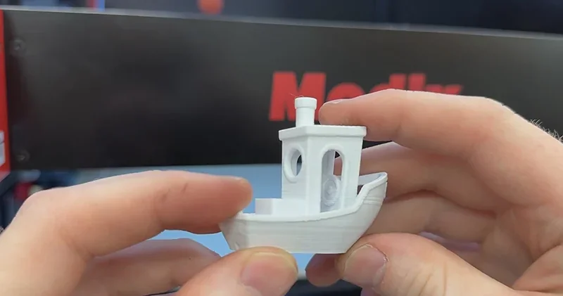 Hands hold a white 3D-printed boat model by a Modix Big 60 printer with visible branding in the background.