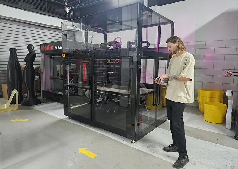 Modix Big 60 3D printer in workshop setting with person operating it, surrounded by large printed sculptures and yellow chairs, showcasing production capabilities.