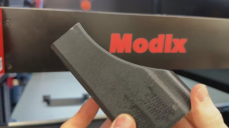 Modix Big 60 3D printer brand logo with a hand holding a printed black part, showcasing the printer's quality and detail.