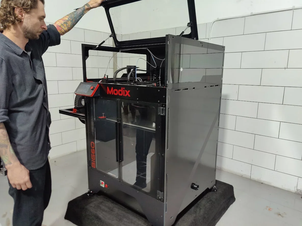 Modix Big 60 3D printer with glass doors, open top, and control panel, in a tiled room. A person interacts with the machine.
