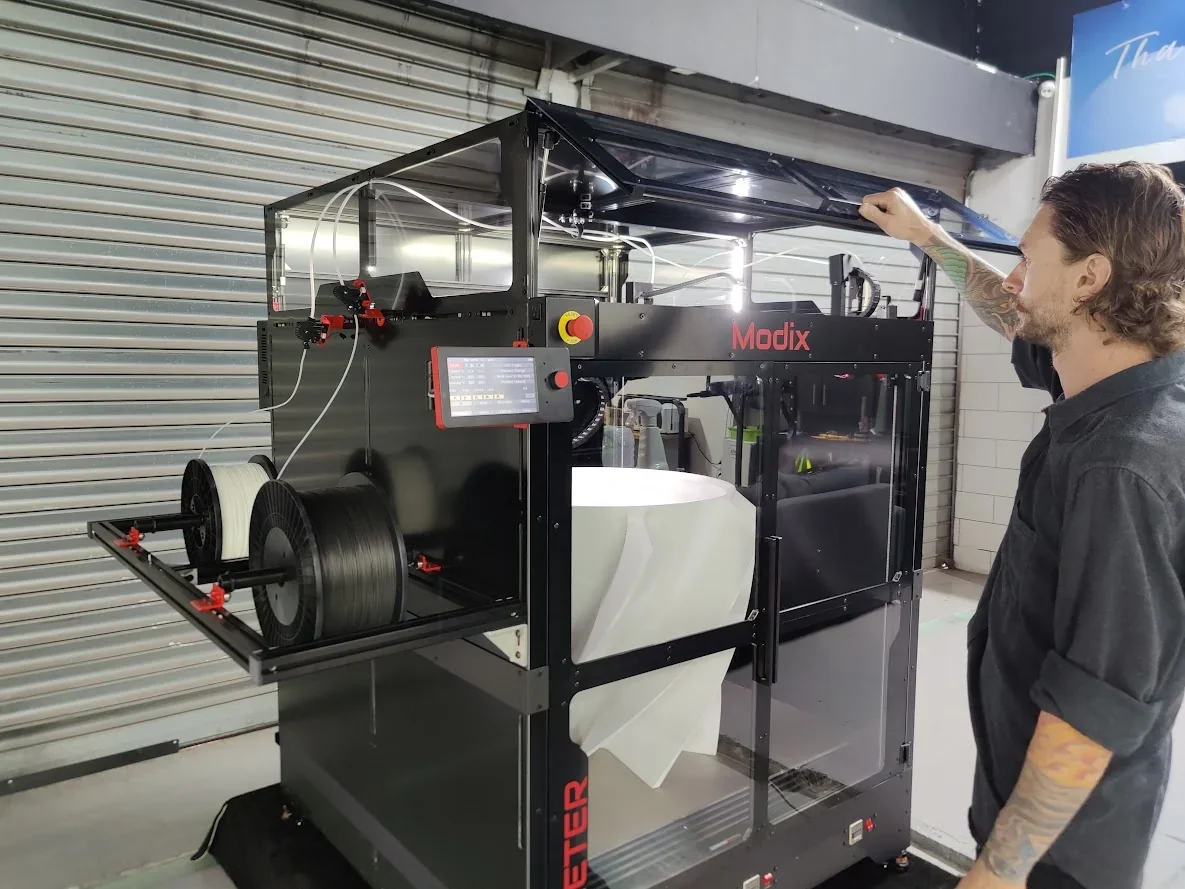 Modix Big 60 3D printer in a workshop setting with filament spools and a person inspecting its transparent enclosure, branding visible.