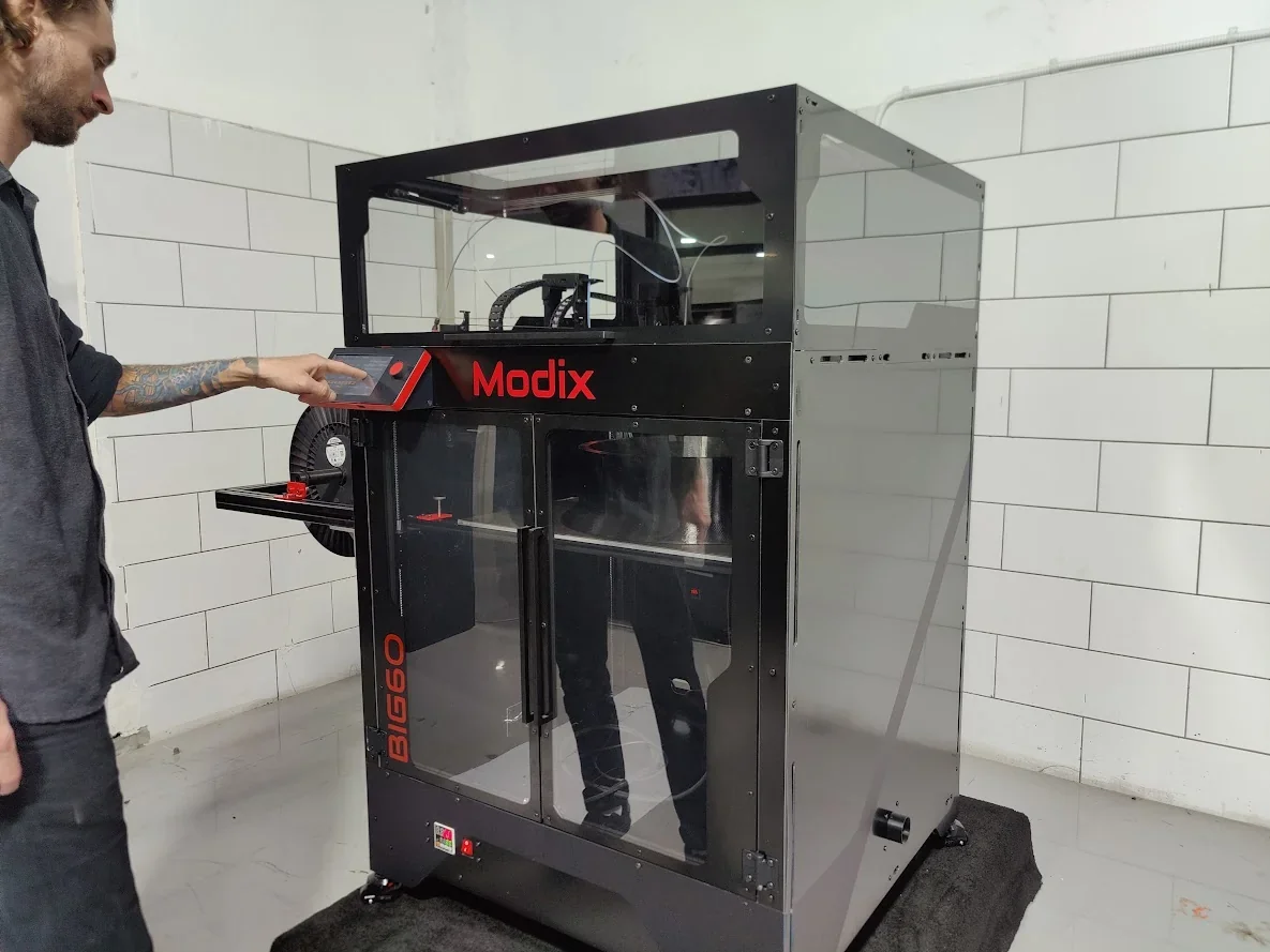 Modix Big 60 3D printer in a tiled room with a person interacting, featuring a visible filament spool and branding.