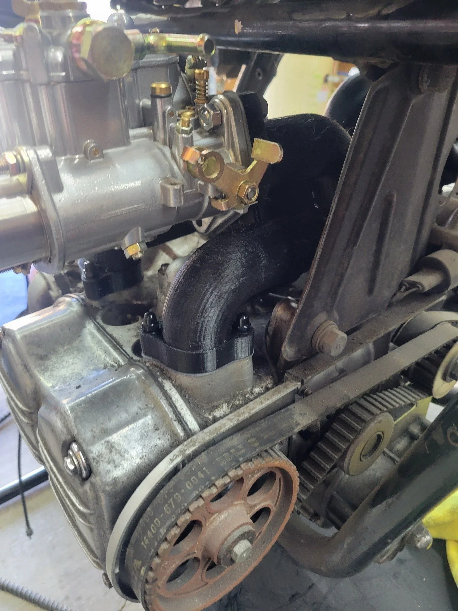 Close-up of an engine with a black 3D printed component created by Modix Big 60, featuring metal gears, belts, and visible mechanical parts.