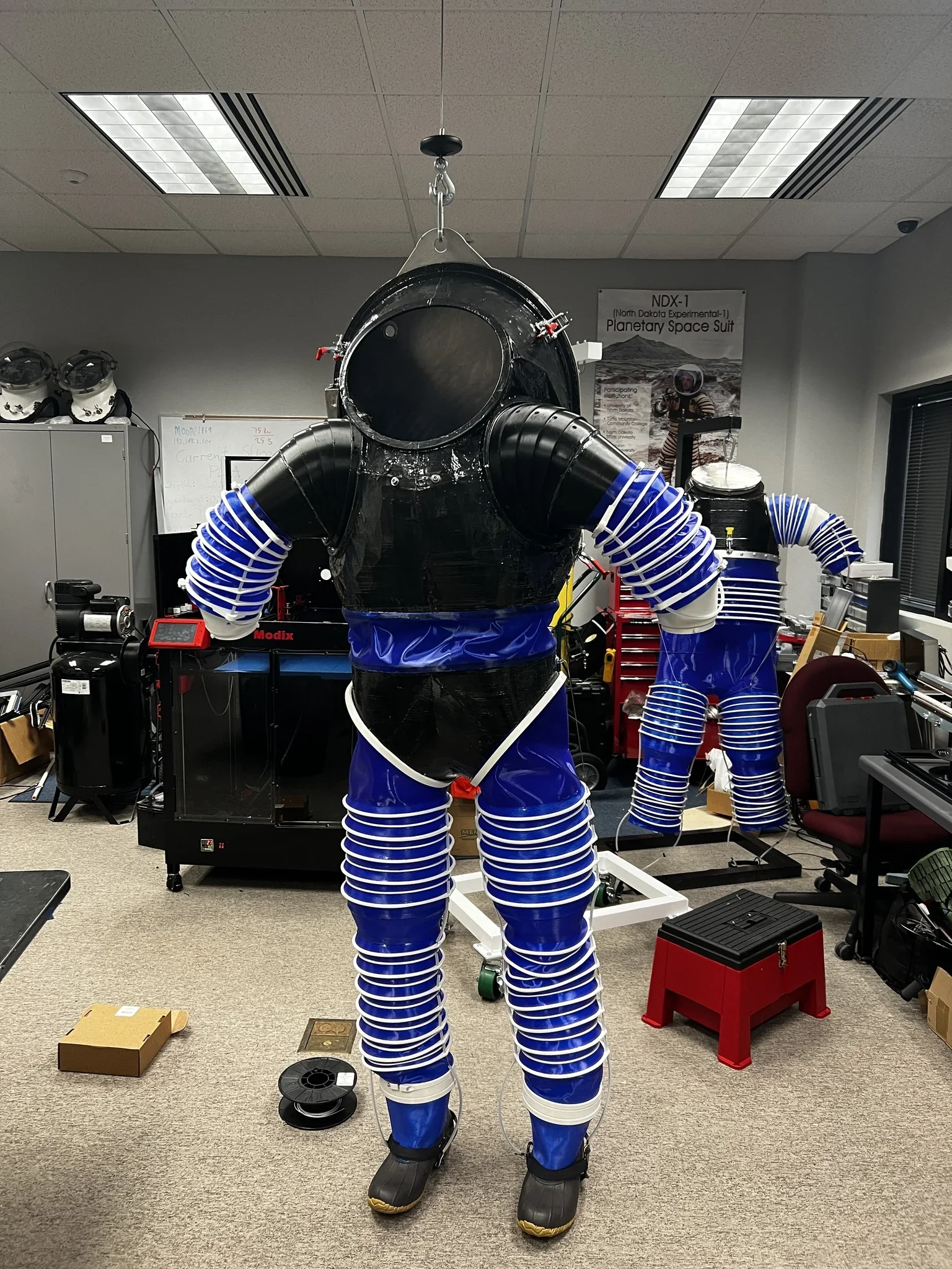 Modix Big Meter 3D printer creating futuristic space suits in a workshop setting, showcasing blue materials with visible branding in the background.