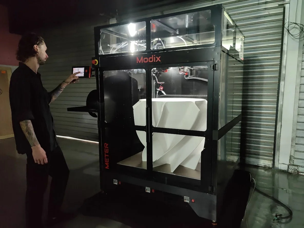 Modix Big Meter 3D printer in a garage setting, printing a large white part, with a person using the control panel.