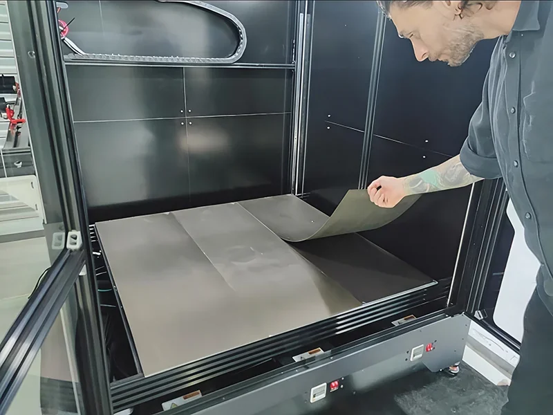 Modix Big Meter 3D printer with open chamber, a person adjusts the flexible metal print bed, showcasing large build volume and machine operations.