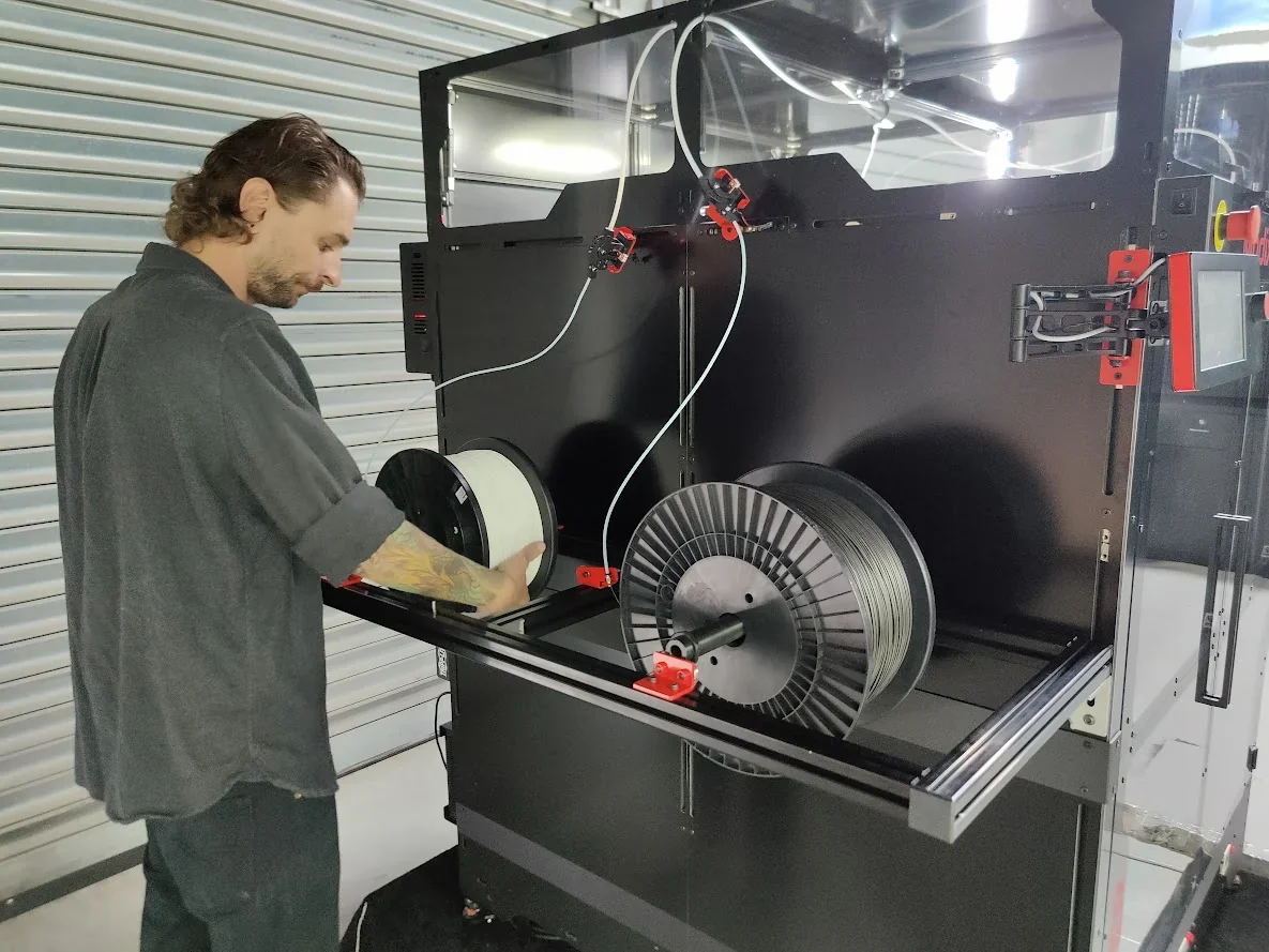 Person adjusting filament spools on Modix Big Meter 3D printer inside a workshop, with metal shutters in the background.