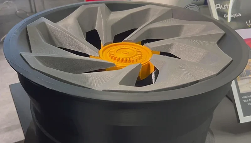 3D-printed wheel with gray spokes and a yellow center, produced by Modix Big Meter, displayed on a table with product info in the background.