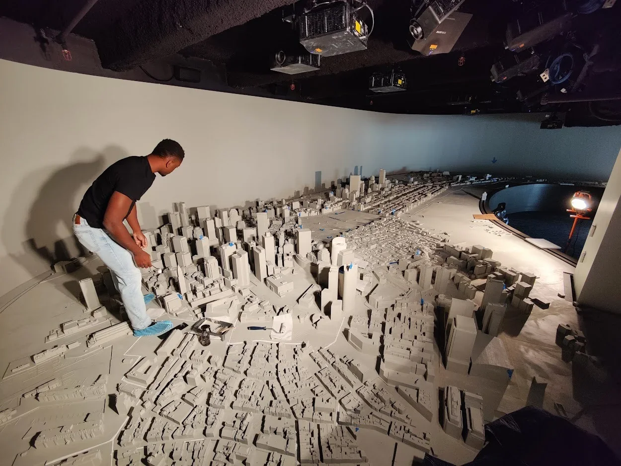 A person inspects a large cityscape model on a table, likely created by the Modix Big Meter 3D printer, under ceiling-mounted projectors.