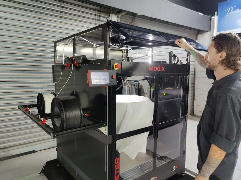 Modix Big Meter 3D printer in industrial setting with operator, large filament spools, and control panel visible.