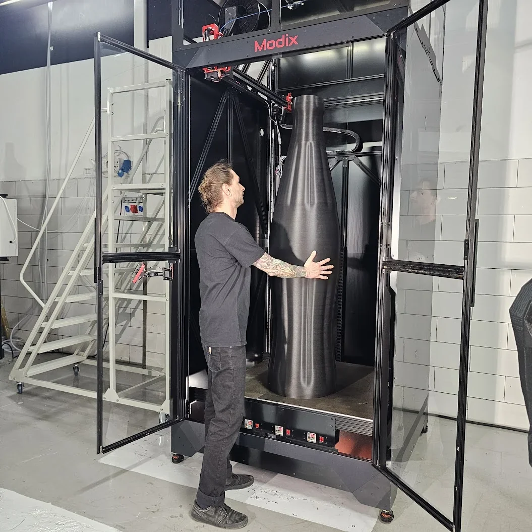 Modix Everest 3D printer with a large printed part inside, man adjusting the model. Black metal frame, industrial setting with visible branding.