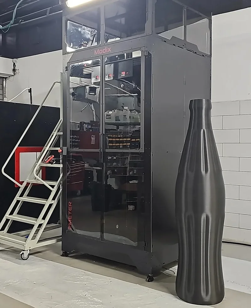 Modix Everest 3D printer with a large printed part beside it, set in a workshop environment, showcasing its size and capability.