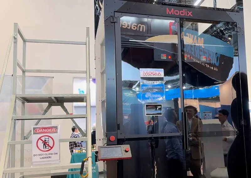 Modix Everest 3D printer at expo with glass front, branding visible. Ladder with warning sign nearby.
