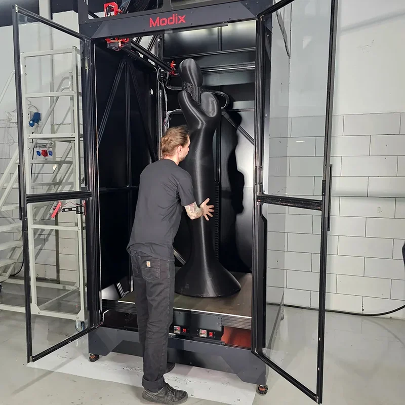 Modix Everest 3D printer with a large black sculpture inside, featuring open doors and a person interacting with the machine in an industrial setting.